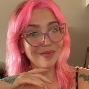A woman with pink hair and striking, cat-eye glasses smiles at the viewer.