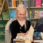 A woman with long blond hair and a black top looking at books while sitting in front of a full bookshelf. 