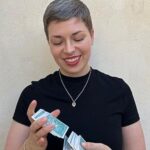 A smiling woman with short hair wearing a black t-shirt and a gold necklace - she is shuffling tarot cards and looking at the cards.