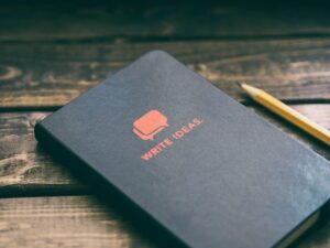 A black notebook with the text “Write Ideas” on it in red, on a wooden surface next to a pencil.