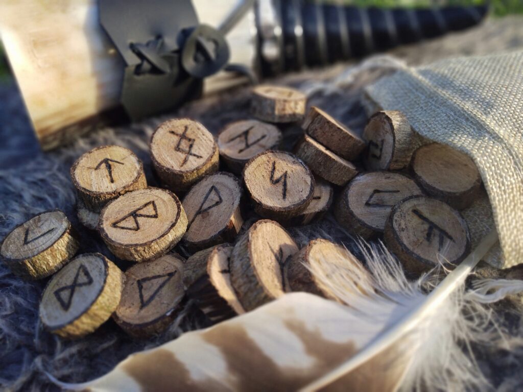 A pile of wooden elder futhark runes spills out of a pale fabric bag onto a dark table; there is a buzzard feather next to the bag.