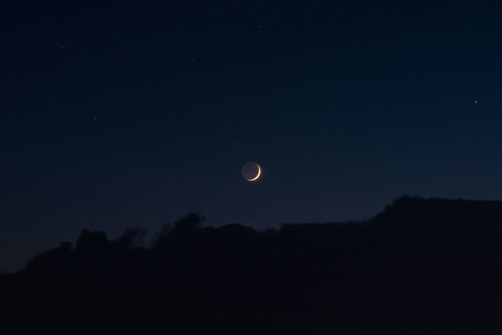 A new moon against a dark blue sky above silhouetted hills.