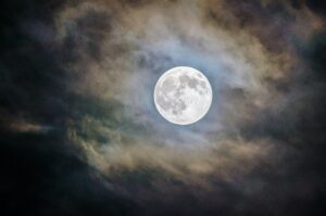 A full moon, casting silvery light on a thin layer of surrounding cloud against a dark sky.